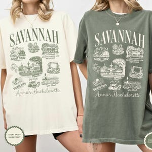 Pu&ograve; includere: Due magliette, una avorio e una verde muschio, ciascuna con un design grafico. Il design include la parola "SAVANNAH" sopra illustrazioni di monumenti e il testo "Anna's Bachelorette". La maglietta avorio ha una stampa muschio.