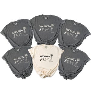 May include: Five short-sleeved t-shirts arranged in a group. Four shirts are dark charcoal gray with white ink, and one center shirt is cream with black ink. Each features the text Wild Wild Crew or Wild Wild Wife, with illustrations of a champagne bottle, a ring, two martini glasses, and a sun.