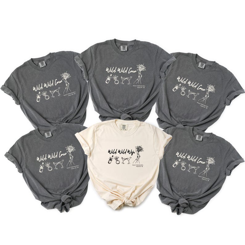 May include: Five short-sleeved t-shirts arranged in a group. Four shirts are dark charcoal gray with white ink, and one center shirt is cream with black ink. Each features the text Wild Wild Crew or Wild Wild Wife, with illustrations of a champagne bottle, a ring, two martini glasses, and a sun.