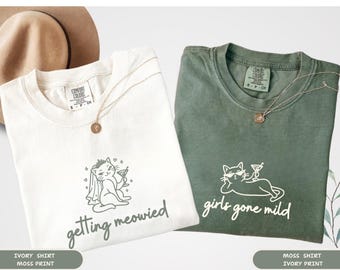 Puede incluir: Dos camisetas, una marfil y otra verde musgo, cada una con un gráfico de gato y texto. La camiseta marfil dice "getting meowied" y la camiseta verde dice "girls gone mild". Ambas camisetas tienen un collar dorado.