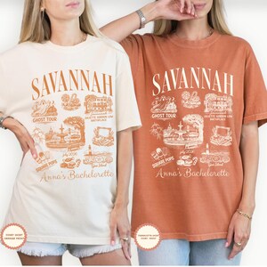 Pu&ograve; includere: Due magliette, una avorio e una terracotta, con una grafica arancione dettagliata. La grafica include la parola "SAVANNAH" sopra illustrazioni di monumenti locali e il testo "Anna's Bachelorette" sotto. Le magliette sono a maniche corte.