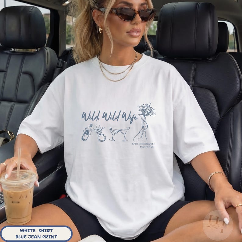 May include: A white short-sleeved t-shirt featuring a blue graphic print with the text Wild, Wild Wife, an illustration of a champagne bottle, a diamond ring, two martini glasses, and a disco ball held by a hand, with Grace's Bachelorette Nashville '26.