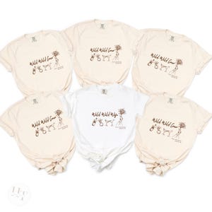 May include: Five cream-colored t-shirts and one white t-shirt featuring a brown line-art graphic. The design includes the text 'Wild Wild Crew' or 'Wild Wild Wife' alongside illustrations of a champagne bottle, a ring, a cocktail glass, and a sunflower.