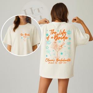 May include: Off-white t-shirt featuring the text "The Bride" on the front and "The Life of a Bride" on the back, with a cocktail glass design. The back also displays "Olivia's Bachelorette" and "Miami, FL EST 2025".