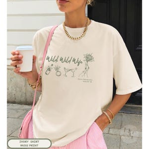 May include: An ivory short-sleeved t-shirt featuring a green graphic print with the text Wild, Wild Wife, a champagne bottle, a diamond ring, two martini glasses, a disco ball, and the text Gracie's Bachelorette Nashville '26.