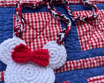 May include: A white crochet purse shaped like a mouse head with a red bow. The purse has a braided strap in red, black, and white. The background is a red and white checkered pattern on a blue quilt.
