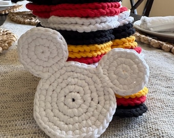 May include: A stack of handmade crochet coasters in a variety of colors, including white, black, red, and yellow. One white coaster is shaped like a mouse head with two round ears. The coasters are stacked on a textured surface.