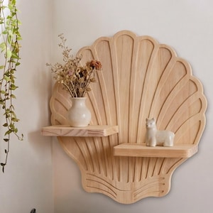Könnte beinhalten: Hellfarbenes Wandregal in Muschelform aus Holz mit zwei kleinen Ablagen. Auf den Ablagen stehen eine kleine weiße Vase mit Trockenblumen und eine weiße Keramikfigur. Das Regal ist an einer weißen Wand befestigt.