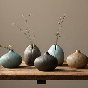 Op de afbeelding: Vijf keramische vazen in aardetinten, waaronder blauw, bruin en beige, staan op een houten oppervlak. Elke vaas heeft een unieke vorm en textuur, sommige met verticale groeven. Delicate gedroogde stelen en bloemen zijn in sommige vazen geplaatst.