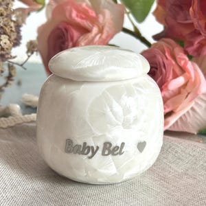 May include: A white, pearlescent urn with a lid, inscribed with "Baby Bel" and a heart symbol. The rounded urn is set against a backdrop of pink roses and greenery, suggesting a memorial theme. The urn is likely for ashes.