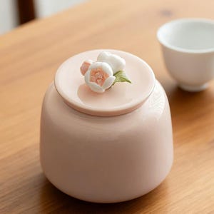 Op de afbeelding: Een lichtroze keramische pot met een bijpassende deksel. De deksel is versierd met een cluster van witte en roze bloesems en groene bladeren. Een witte theekop staat op de achtergrond. De pot staat op een houten oppervlak.