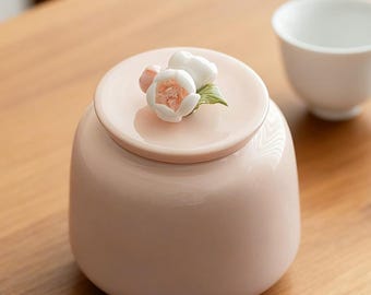 May include: A pale pink ceramic jar with a matching lid. The lid is adorned with a cluster of white and pink flower blossoms and green leaves. A white teacup is in the background. The jar is on a wooden surface.
