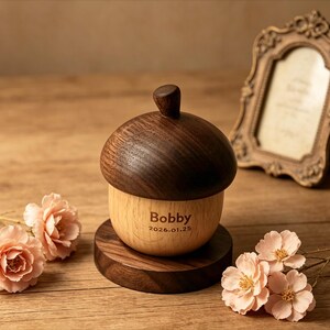 May include: A wooden acorn-shaped keepsake box. The top is dark brown, the base is light brown, and it's engraved with "Bobby" and "2026.01.25". It sits on a round wooden stand. A decorative picture frame and pink flowers are in the background.