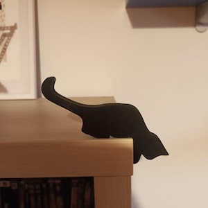 May include: A black cat-shaped shelf sitter, crafted from a solid material, is perched on the edge of a wooden shelf. The silhouette of the cat is sleek and modern, adding a touch of whimsy to the decor. The shelf sitter is a decorative item.