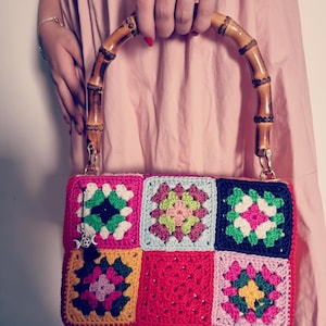 May include: A colorful, handmade crochet bag with a bamboo handle. The bag features a patchwork design of squares in various colors, including pink, red, green, and yellow. The handle is made of curved bamboo, and the bag has gold-colored hardware.
