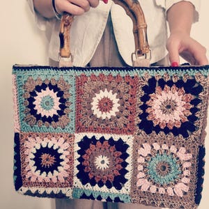 May include: A handmade crochet bag with a bamboo handle. The bag features a patchwork design of colorful squares in shades of blue, pink, brown, and cream. The bamboo handle is curved and has a natural wood grain finish. The bag is held by a person.