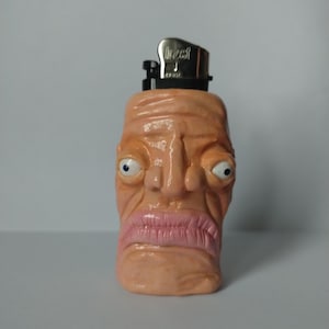 Könnte beinhalten: Ein neuartiges Feuerzeug in Form eines menschlichen Gesichts. Das Feuerzeug hat ein pfirsichfarbenes Gesicht mit übertriebenen Gesichtszügen, darunter große Augen, eine markante Nase und rosa Lippen. Die Oberseite des Feuerzeugs ist silberfarben.