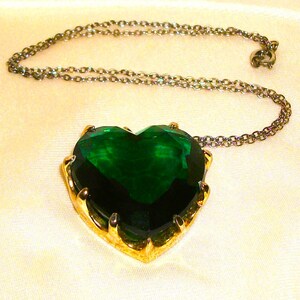 May include: A heart-shaped pendant necklace with a large, emerald-green gemstone. The gemstone is set in a gold-colored metal frame and hangs from a delicate silver-colored chain. The necklace is displayed on a cream-colored, textured fabric.