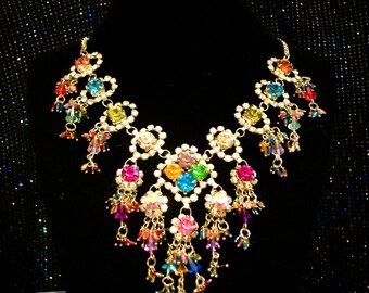 May include: A gold-tone statement necklace featuring a central cluster of multi-colored rhinestones in pink, orange, green, and blue. The piece is adorned with dangling crystal beads in various hues and clear rhinestones set along the metal frame.