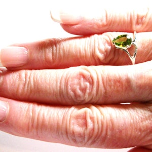 May include: A close-up of a hand wearing a silver ring with a pear-shaped green gemstone. The ring is on the middle finger of the hand.