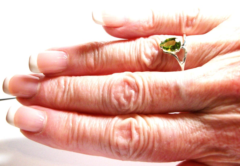 May include: A close-up of a hand wearing a silver ring with a pear-shaped green gemstone. The ring is on the middle finger of the hand.