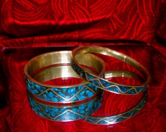 May include: Two metal bangles featuring a gold-toned finish and inlaid blue stone fragments. The bracelets are decorated with a repeating zigzag pattern that separates the crushed blue stone inlay, creating a distinct geometric design on each band.