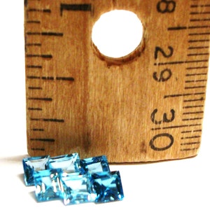 May include: A group of six blue topaz gemstones, each measuring approximately 1/2 inch in length, are arranged on a white surface next to a wooden ruler. The ruler is marked with inches and has a hole in the center.