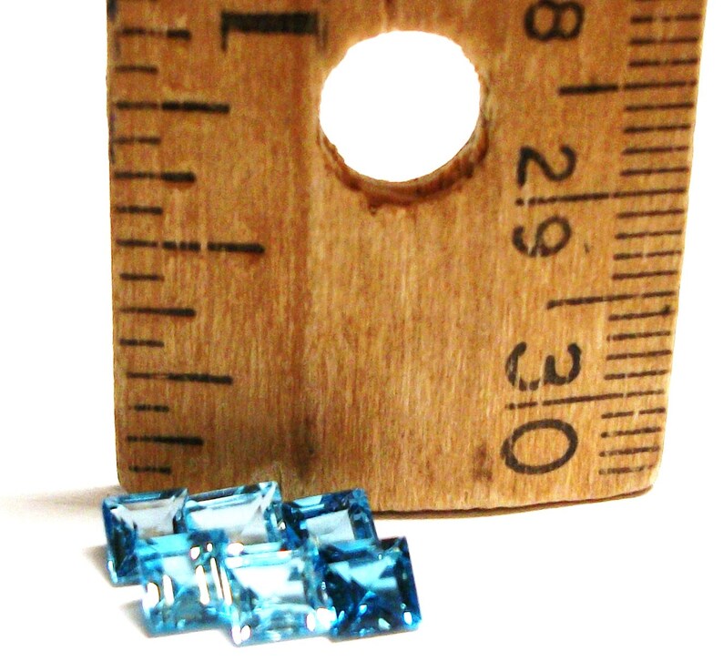 May include: A group of six blue topaz gemstones, each measuring approximately 1/2 inch in length, are arranged on a white surface next to a wooden ruler. The ruler is marked with inches and has a hole in the center.