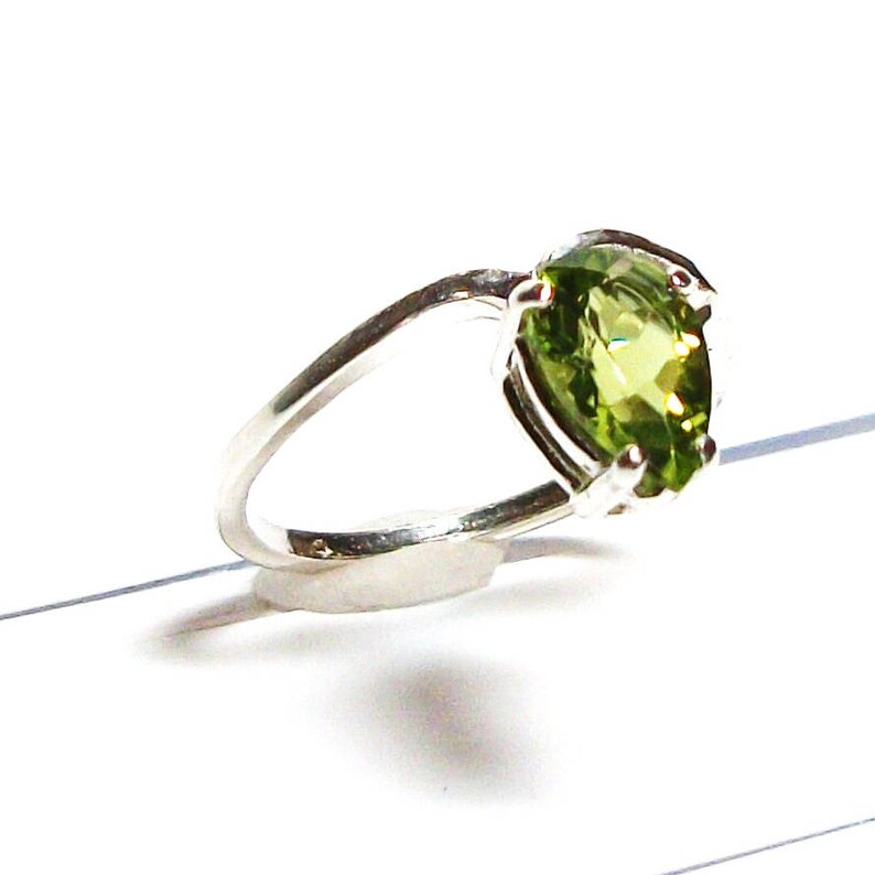 May include: A silver ring featuring a large, oval-shaped green gemstone. The ring is set with small, clear stones around the gemstone.