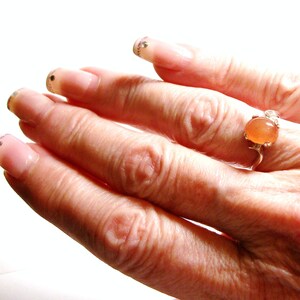 May include: A hand wearing a ring with a light orange oval gemstone. The ring is made of silver and has a delicate design. The hand is wearing clear nail polish with glitter accents.