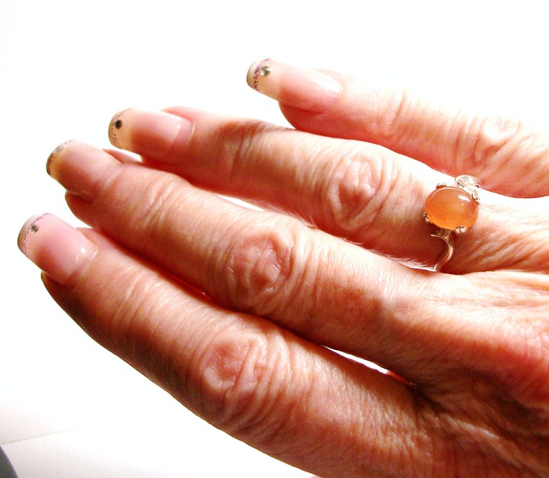 May include: A hand wearing a ring with a light orange oval gemstone. The ring is made of silver and has a delicate design. The hand is wearing clear nail polish with glitter accents.