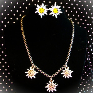 May include: A jewelry set consisting of a necklace and matching stud earrings. The necklace features a metallic chain with three edelweiss flower charms. The earrings are shaped like individual white and yellow edelweiss flowers.