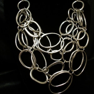 May include: A silver-toned statement necklace featuring a cascade of interconnected oval and circular links. The necklace has a bold, chunky design with varying sizes of links, creating a layered effect. The metal has a polished finish, reflecting light and adding a touch of glamour.