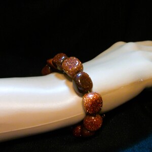 May include: A beaded bracelet featuring large, rounded, reddish-brown stones with a sparkling, glittery appearance. The bracelet is displayed on a mannequin arm against a dark background, showcasing the jewelry's color and texture.