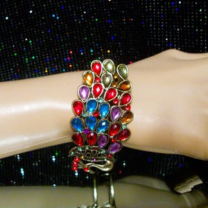 May include: A gold-toned bracelet with a peacock design, featuring colorful teardrop-shaped gemstones in red, blue, purple, and yellow. The bracelet is displayed on a mannequin arm against a sparkly black background.