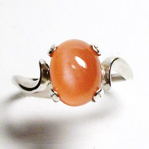 May include: A silver ring with a peach-colored oval gemstone set in a prong setting. The ring band is curved and has a smooth finish.