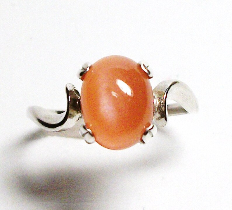 May include: A silver ring with a peach-colored oval gemstone set in a prong setting. The ring band is curved and has a smooth finish.