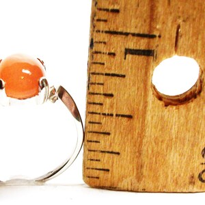 May include: A silver ring with a large, round, orange gemstone. The ring is next to a ruler, showing the size of the stone.