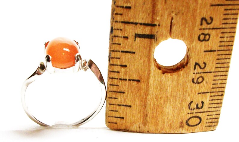 May include: A silver ring with a large, round, orange gemstone. The ring is next to a ruler, showing the size of the stone.