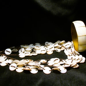 May include: A layered necklace and a bracelet. The necklace features multiple strands of small, white, circular beads with dark accents. The bracelet is a wide band with a gold interior and white, rectangular segments.
