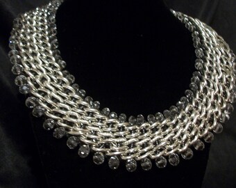 May include: A silver-toned chain necklace with multiple rows of interlocking links. The necklace is adorned with small, dark gray beads along the bottom edge. The jewelry is displayed on a black mannequin.