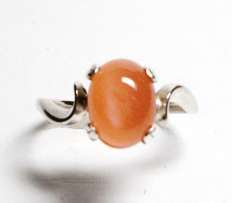 May include: A silver ring with a peach-colored oval gemstone set in a prong setting. The ring band is curved and has a smooth finish.