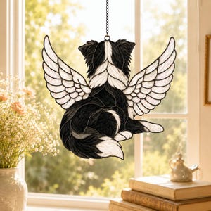 May include: A stained glass suncatcher depicting a black and white dog with large, feathered angel wings. The dog is shown from behind, featuring textured black and white glass pieces joined by dark metal solder lines, suspended by a metal chain.