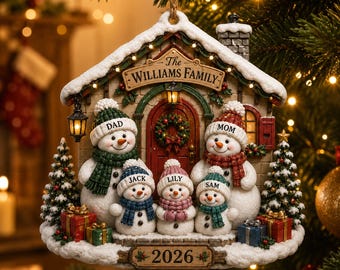 May include: A house-shaped Christmas ornament featuring five snowmen wearing hats and scarves labelled Dad, Mom, Jack, Lily, and Sam. The structure displays the text The Williams Family and 2026, decorated with miniature trees, gifts, and lights.