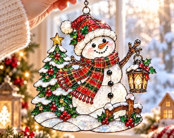 May include: A stained glass-style hanging ornament featuring a snowman wearing a red and green plaid scarf and a matching hat. The snowman stands next to a decorated Christmas tree and holds a glowing lantern, all accented with holly berries.