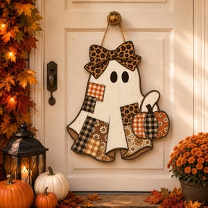 May include: A white ghost-shaped wooden door hanger featuring a leopard print bow, patchwork patterns in orange, black, and white plaid, and floral designs. The ghost holds a small pumpkin with orange and black plaid sections, suspended by a rope.