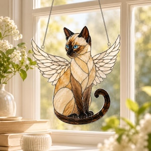 May include: A stained glass suncatcher depicting a Siamese cat with large white feathered wings. The cat features tan and dark brown glass segments, bright blue eyes, and a curled tail. It is suspended by two thin metal chains from a window frame.