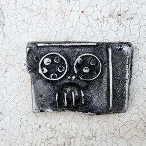 Peut inclure: Une sculpture métallique rectangulaire en forme de robot avec une finition vieillie. La sculpture présente deux détails circulaires en forme d'yeux avec de plus petits cercles à l'intérieur, une forme de bouche et des lignes verticales. La couleur est principalement gris foncé avec des reflets argentés.