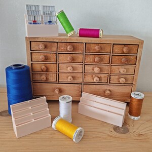 Op de afbeelding: Een houten naaibox met meerdere kleine lades, met spoelen garen in verschillende kleuren, waaronder blauw, groen, roze, geel, wit en bruin. Naaimachinenaalden in een witte houder. Een blauwe garenklos aan de linkerkant.