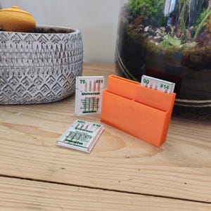 Peut inclure: Un support orange en plastique pour aiguilles à coudre avec trois paquets d'aiguilles. Les paquets sont étiquetés "Universal", "Glamour", et ont des tailles d'aiguilles #11, #16 et #14. Le support est sur une surface en bois.